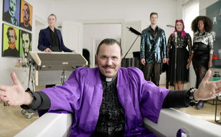 Orphea in Love, Szenenbild, frontal: Ein Mann mit Schnurrbart und bekleidet mit einem lilafarbenen Predigerumhang sitzt in einer Badewanne und breitet lächelnd die Hände aus. Im Hintergrund sind ein dreiköpfiger Chor und ein Keyboarder zu sehen. (© missingFILMs)