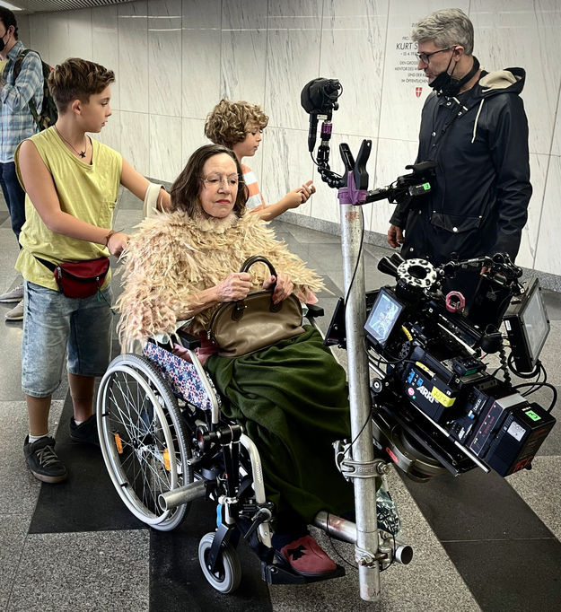 Setfoto vom Dreh zu Neue Geschichten vom Franz: Die Darstellerin von Frau Berger sitzt wartend in einem Rollstuhl, an dem vorne ein Statuv mit Kamera angebracht ist (© Matthias Grunsky)