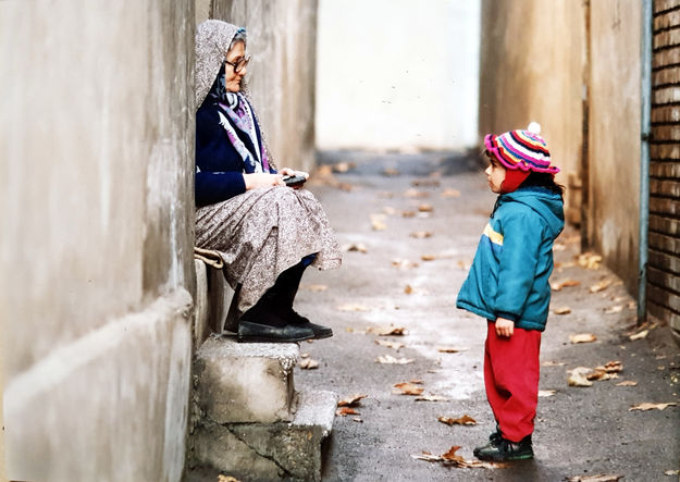 Ein Sack Reis, Szenenbild: In einer engen Gasse sitzt eine alte Frau am linken Bildrand auf der Stufe einer Tür. Ihr gegenüber steht ein Kind. Beide schauen sich an. (Foto: Mitra Mahasani, Produzent: Mohammad Ali Talebi. © Mohammad Ali Talebi)