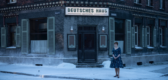 Deutsches Haus, Szenenbild: Eine junge Frau in einem Kleid der 1960er-Jahre steht auf einem schneebedeckten Gehweg vor einer Gaststätte, die "Deutsches Haus" heißt. Die Szenerie ist in blau-grauen Farbtönen gehalten. (© 2023 Disney+)