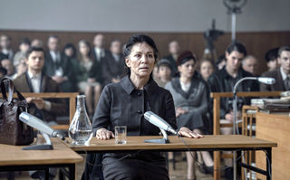 Deutsches Haus, Szenenbild: Eine ältere Frau in einem grauen Kostüm sitzt als Zeugin an einem Tisch in einem Gerichtssaal. Im Hintergrund sind Zuhörer/-innen zu sehen. (© 2023 Disney+)