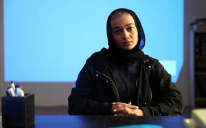 Szenenbild aus dem Film "Irdische Verse": Eine junge, schwarz gekleidete Frau sitzt in einem büroartigen Raum vor einer blau beleuchteten Wand. Sie hat kurze blonde Haare, die sie mit einem dunklem Tuch bedeckt hat. Sie schaut direkt in die Kamera. (© Neue Visionen Filmverleih)