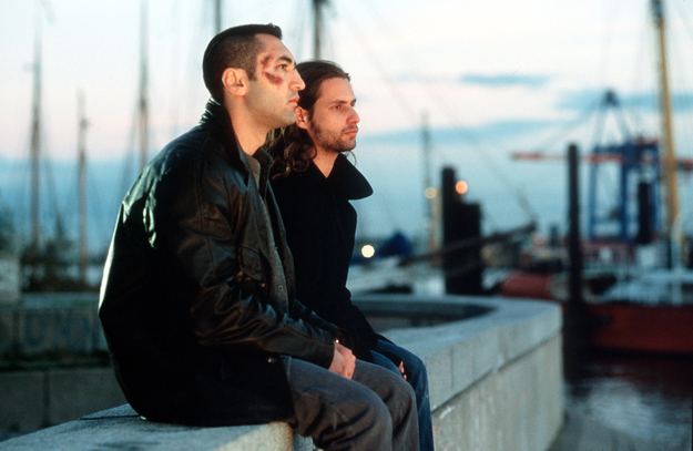 Kurz und schmerzlos, Szenenbild: Zwei junge Männer sitzen nebeneinander auf einer Mauer. Der eine Mann ist im Gesicht verletzt. Im Hintergrund sind Segelboote zu erkennen. (© picture alliance/United Archives | United Archives / kpa Publicity)