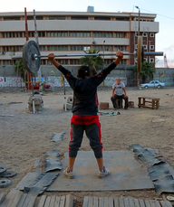 Lift Like a Girl, Szenenbild: Auf einem staubigen Platz stemmt eine Sportlerin vor den Augen eines älteren Mannes ein Gewicht.(© ZDF/Mayye Zayed)