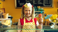 Lucy ist jetzt Gangster, Szenenbild: Ein Mädchen mit Zöpfen und weißer Mütze steht hinter dem Tresen einer Eisdiele und hält lächelnd eine Eistüte in der Hand. (© INDIFilm/Daniel Dornhöfer)