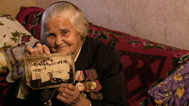 Meine Mutter, ein Krieg und ich, Szenenbild: Eine alte Dame und Veteranin sitzt auf ihrem Sofa und hält lächelnd eine Schwarz-Weiß-Fotografie in die Kamera (© Johann Feindt Filmproduktion)