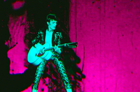 Moonage Daydream, Szenenbild: David Bowie in den 1970ern steht mit einer Gitarre auf einer Bühne. Er selbst ist ein einem grünlichen Ton, der Hintergrund in einem knalligen Pink gehalten. (© UPI Germany)