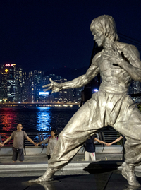 Ein Bild aus Hongkong: Im Vordergrund ist eine Statue des sinoamerikanischen Kampfkünstlers und Schauspielers Bruce Lee zu erkennen. Dahinter stehen Menschen, die sich an den Händen halten und offenbar demonstrieren. Im Hintergrund ist die nächtlichen Skyline von Hongkong zu erkennen.(© Miguel Candela, SOPA Images via ZUMA Wire, picture alliance)
