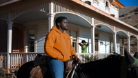 Nope, Szenenbild: Ein Schwarzer Mann im orangenen Pullover sitzt vor einem Haus mit Veranda auf einem Pferd. Im Hintergrund ist eine Schwarze, grün gekleidete Frau zu sehen. (© 2022 Universal Studios. All Rights Reserved)