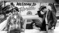 Stranger than Paradise, Szenenbild: Schwarz-weiße Aufnahme von zwei Männern, die an einem Auto stehen. Eine junges Mädchen sitzt hinten im Auto und schaut aus dem geöffneten fenster hinaus. Alle drei tragen Sonnenbrillen. Im Hintergrund steht auf einer Hauswand: Welcome to Florida. (© United Archives /kpa Publicity / Picture Alliance)