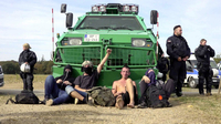 Vergiss Meyn nicht, Szenenbild: Vier teils vermummte Personen sitzen vor einem grünen Räumfahrzeug der Polizei, neben dem uniformierte Beamt/-innen stehen (© W-FILM)