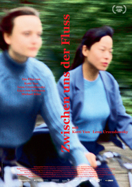 Plakat zum Film "Zwischen uns der Fluss": Bildfüllend sind zwei junge, blau gekleidete Frauen zu sehen, die nebeneinander Fahhrad fahren. Den Hintergrund füllt das Grün von Bäumen oder Büschen aus. Das Bild ist verwischt. Der Filmtitel verläuft vertikal genau zwischen den beiden Frauen in roter Farbe. (© Real Fiction Filmverleih)
