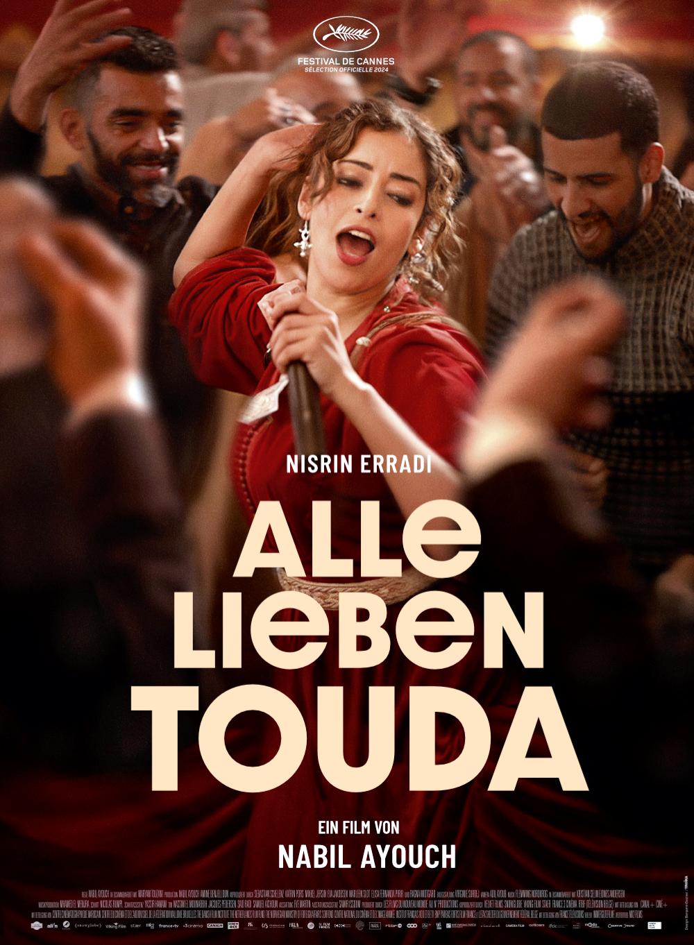 Filmplakat zum Film ALLE LIEBEN TOUDA: Eine junge Frau im roten Kleid singt und tanzt in der Mitte einer Männergruppe. In der unteren Bildhälfte steht der Titel des Films.