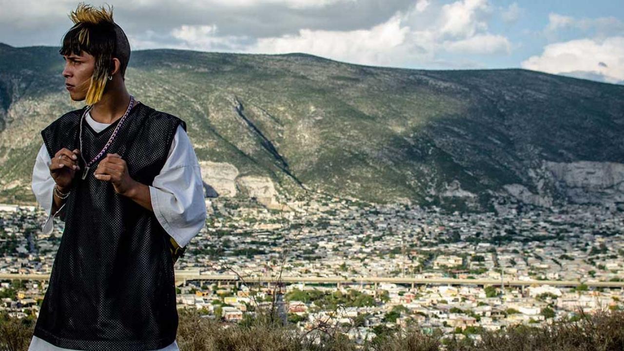 Eine junger Mann steht am linken Rand des Bild, hinter ihm eine Berglandschaft.