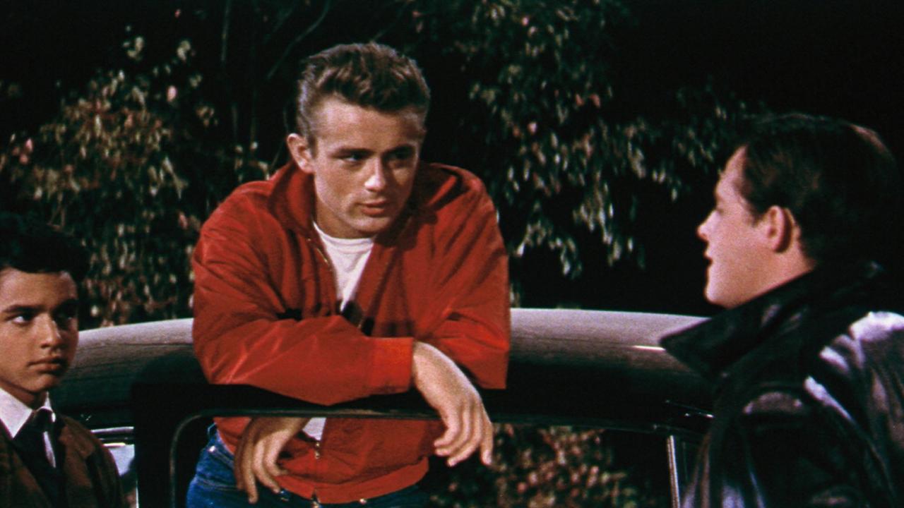 Nachts, ein junger Mann (James Dean) in einem roten Blouson lehnt an einer Autotür und schaut zu einem anderen jungen Mann in schwarzer Lederjacke. Links steht ein junge im braunen Anzug.