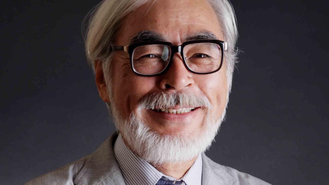 Ein lächelnder Miyazaki Hayao mit weißen Haaren, schwarze Brille und Anzug mit Krawatte. 