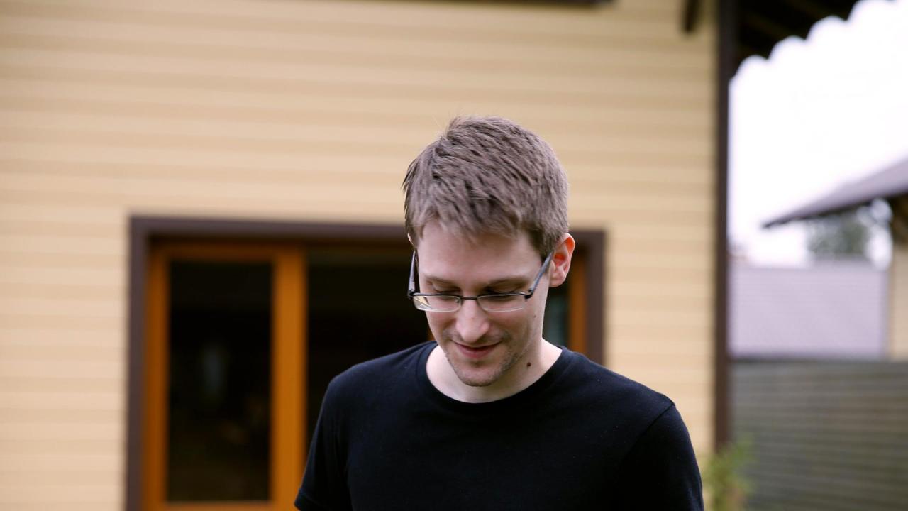 Edward Snowden trägt ein schwarzes T-Shirt und steht vor einem Haus. Es ist eine Nahaufnahme und er guckt auf dem Boden.