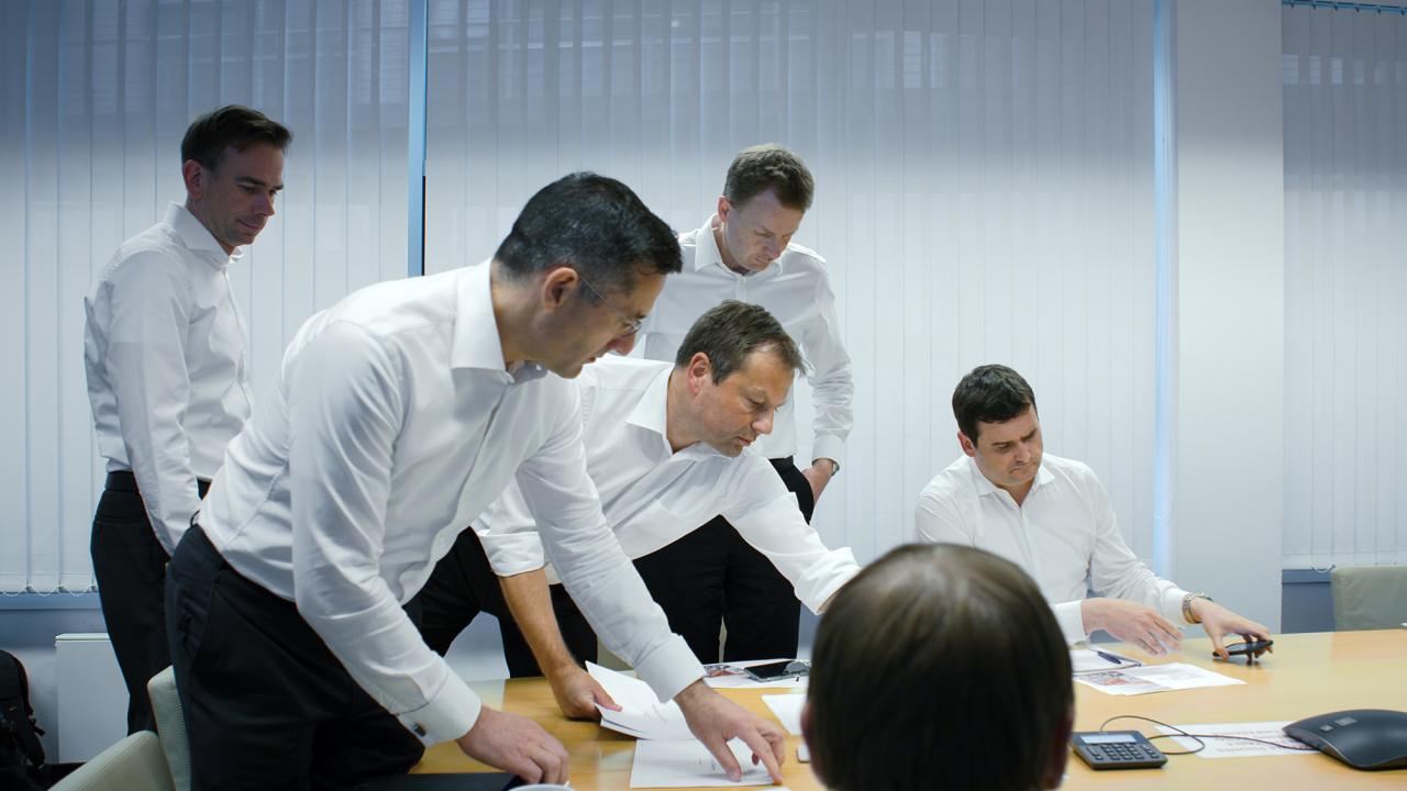 Mehrere Männer in Businesslook umringen einen Bürotisch.