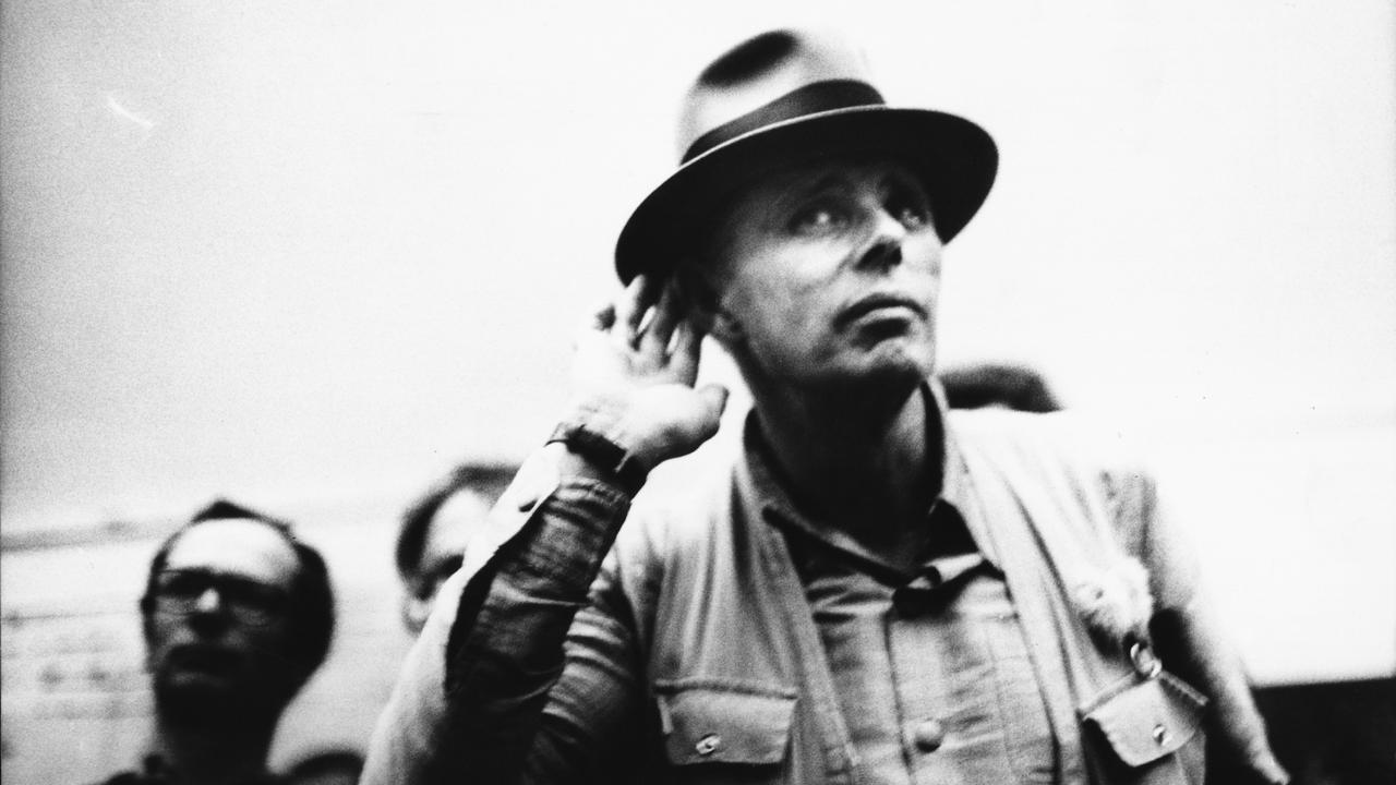 Beuys, Szenenbild: Schwarzweißfoto eines Mannes mit Hut, der die Hand ans Ohr legt, als würde er aufmerksam zuhören.