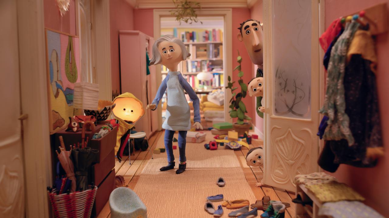 Szenenbild aus dem Puppentrickfilm TONY, SHELLY UND DAS MAGISCHE LICHT: In einem unaufgeräumten Wohnungsflur steht eine Frau. Links von ihr steht ein Junge mit leuchtendem Kopf. Rechts von ihr schauen ein Baby und ein Mann hinter der Tür hervor. Alle blicken direkt in die Kamera.
