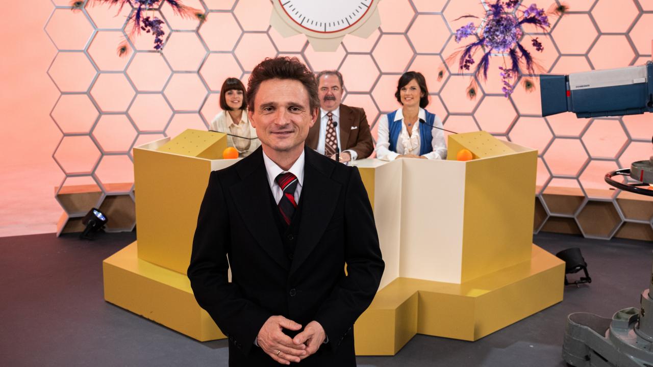 Filmstill aus dem TV-Spielfilm ROSENTHAL: In den 1970er-Jahren steht Hans Rosenthal (Florian Lukas) im schwarzen Anzug vor einer Wabenwand im Studio. Hinter ihm sitzt seine Ratejury, Mady Riehl (Theresia Wald), "Ekki" Fritsch (Timo Dierkes) und Brigitte Xander (Katharina Völkl), an Pulten.