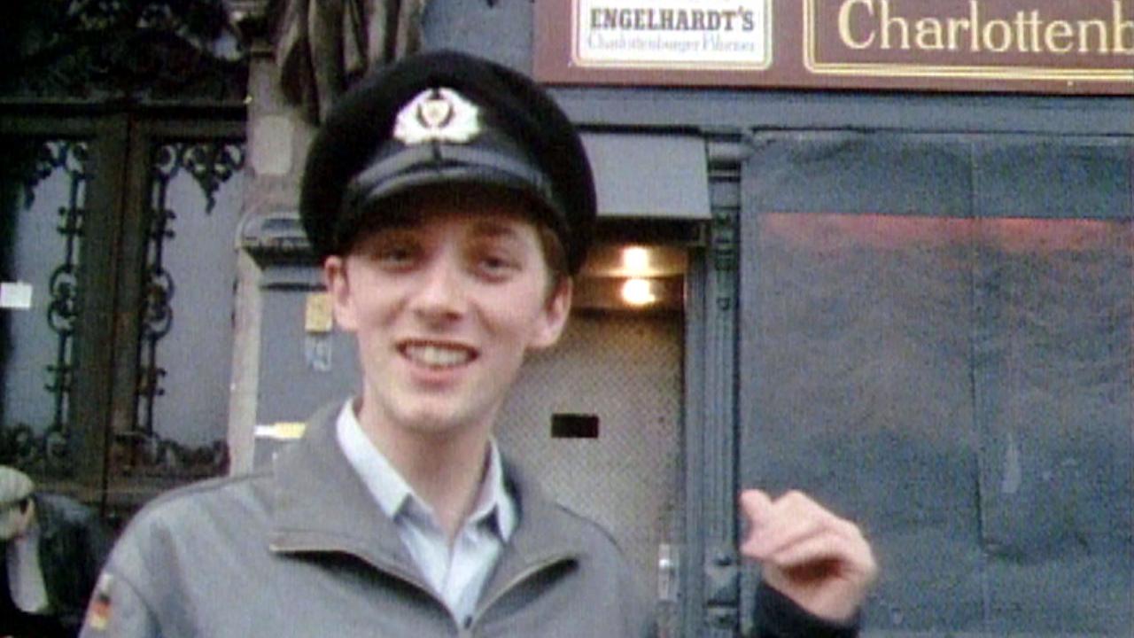 Still aus dem Dokumentarfilm B-MOVIE: LUST & SOUND IN WEST-BERLIN 1979-1989: Ein juger Mann in Militärkappe steht 1985 vor der Eingangstür der Berliner Kneipe Risiko. Er lächelt in die Kamera..