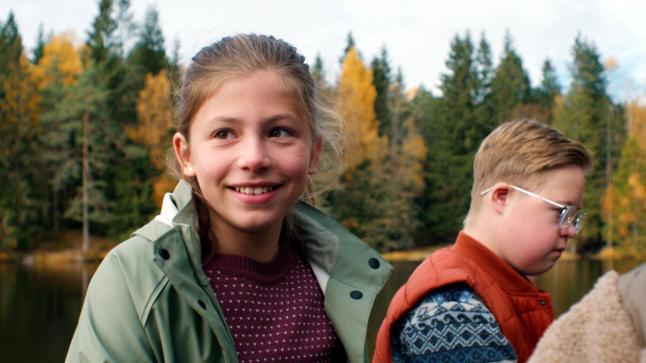 Filmstill aus LARS IST LOL: Bild eines lächelnden Mädchen, neben ihm steht ein Junge mit Down Syndrom; im Hintergrund ist ein herbstlicher Wald zu sehen.