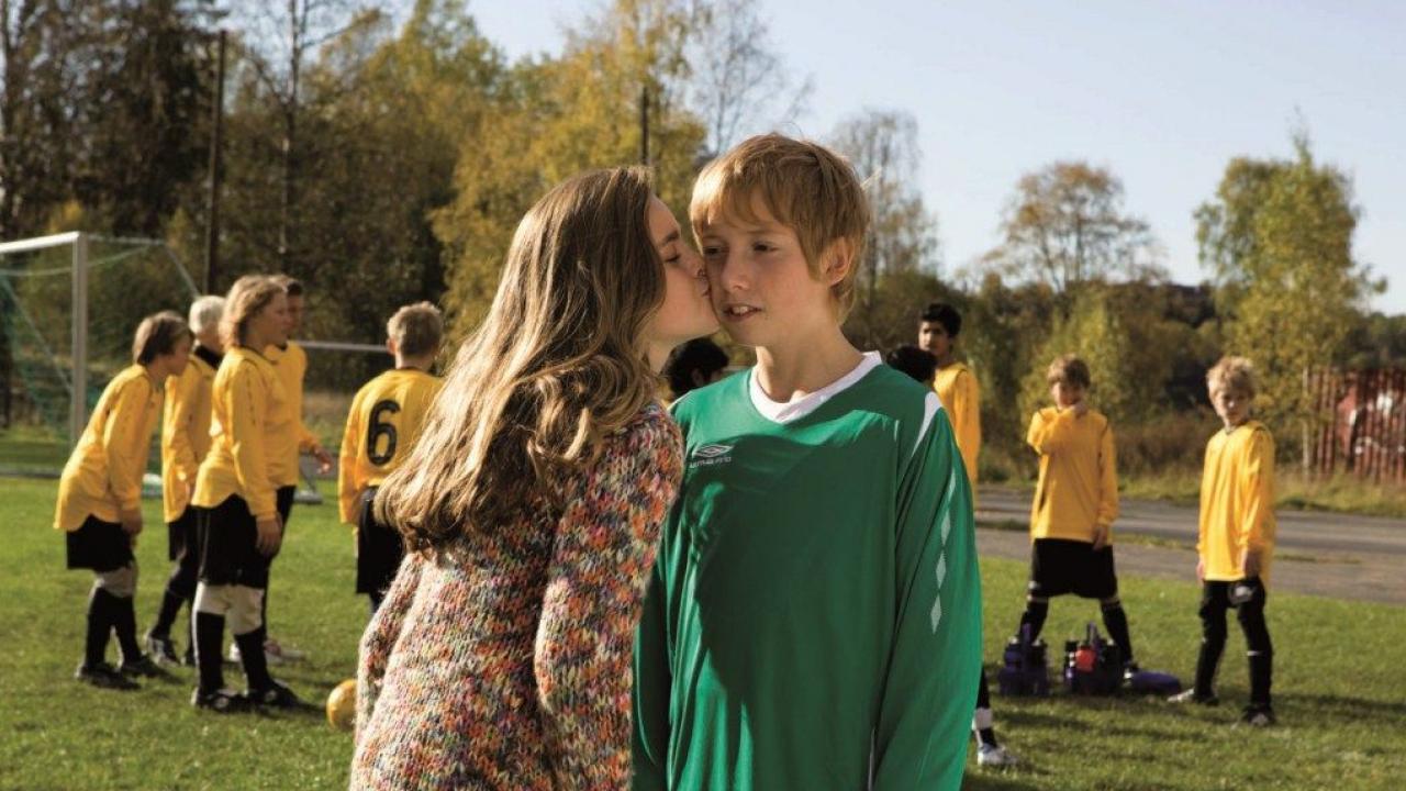 Still aus dem Kinderfilm THE LIVERPOOL GOALIE: Ein Junge im grünen Fußballtrikot bekommt von einem Mädchen einen Kuss auf die Wange. Im Hintergrund sieht man Kinder in Fußballtrikots und einen Fußballplatz.