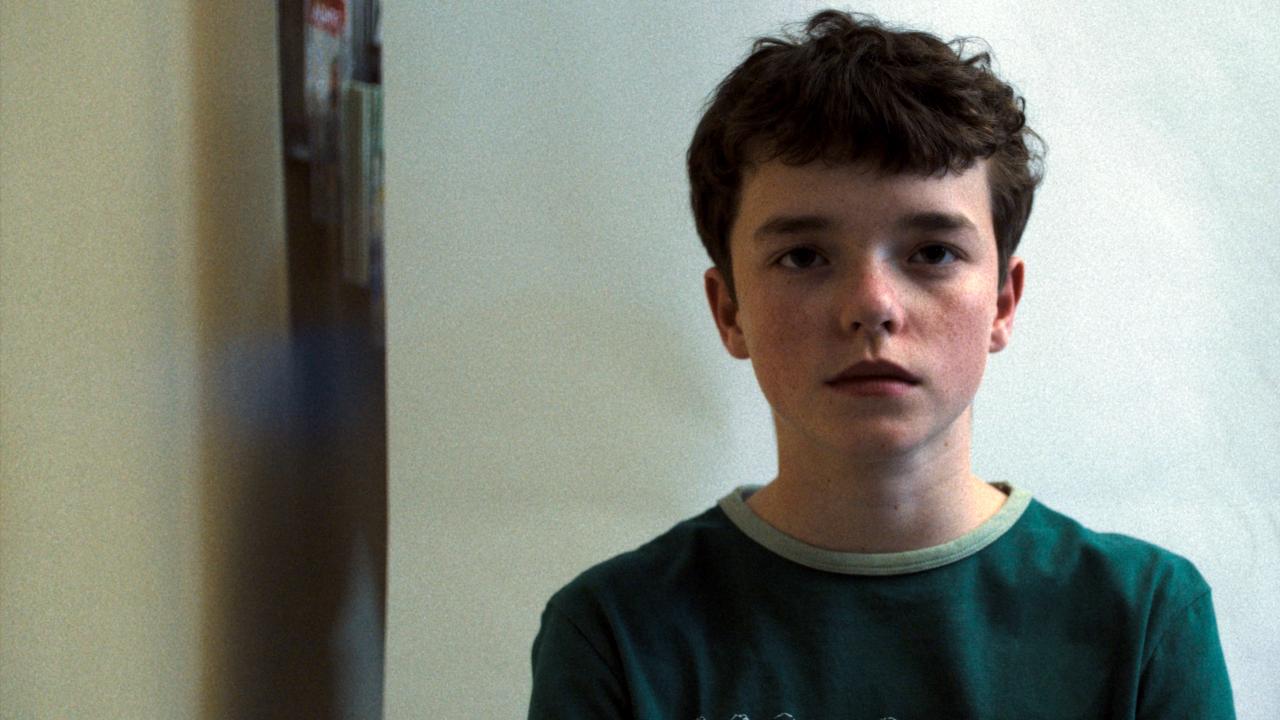 Still aus der Mini-Serie ADOLESCENCE: Porträt eines Jungen im Teenager-Alter, der in einem Büro sitzt und direkt in die Kamera schaut.