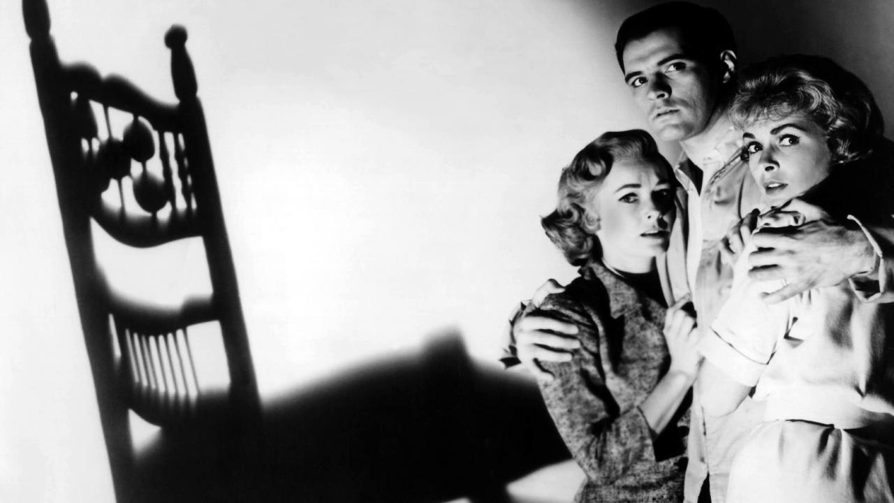 Filmstill aus PSYCHO: Ein Mann hält zwei verängstigte Frauen im Arm.  Die drei stehen am Bildrand, neben ihnen ist der riesige Schatten eines Stuhls zu sehen. 
