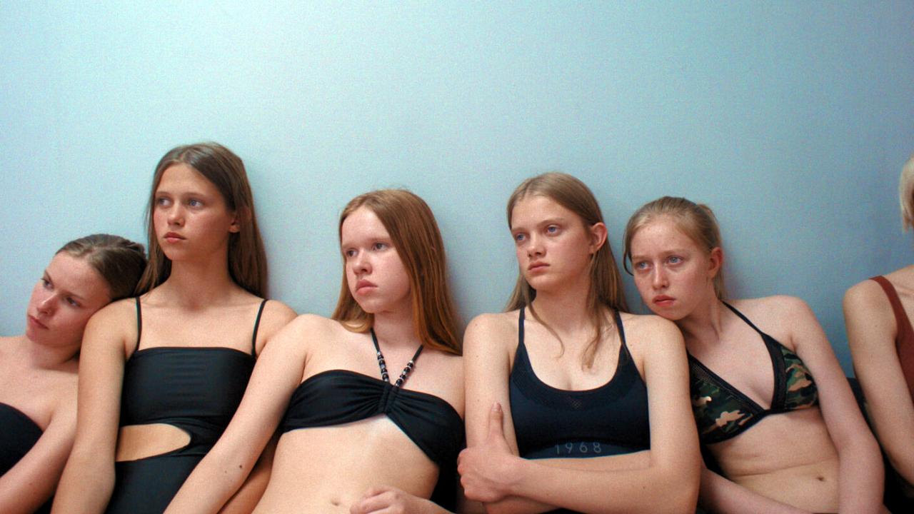 Still aus dem Drama TOXIC: Fünf sehr junge Mädchen, in Bikinis und engen schwarne Bodys bekleidet, sitzen vor einer hellblauen Wand nebeneinander. Ihre Gesichter wirken ausdruckslos.