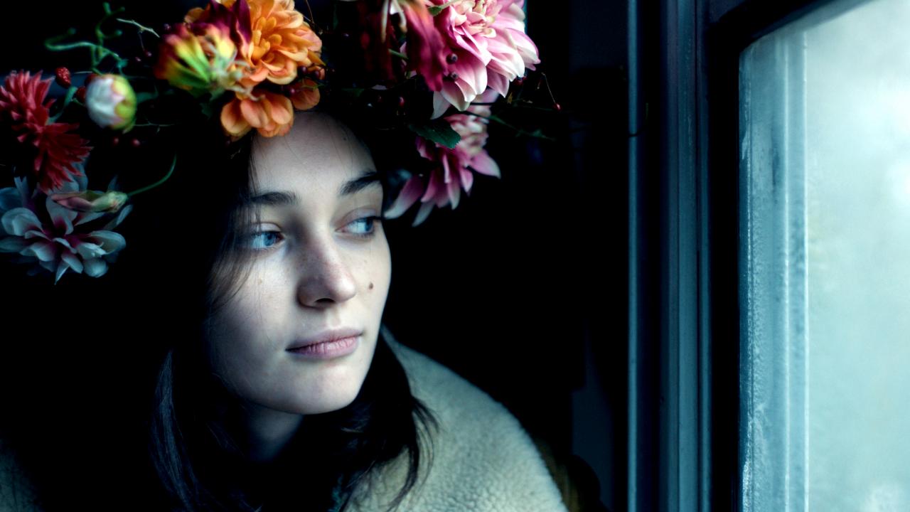 Still aus dem Biopic OXANA: Eine junge Frau schaut aus einem Fenster. Sie trägt einen Blumenkranz auf den Kopf.
