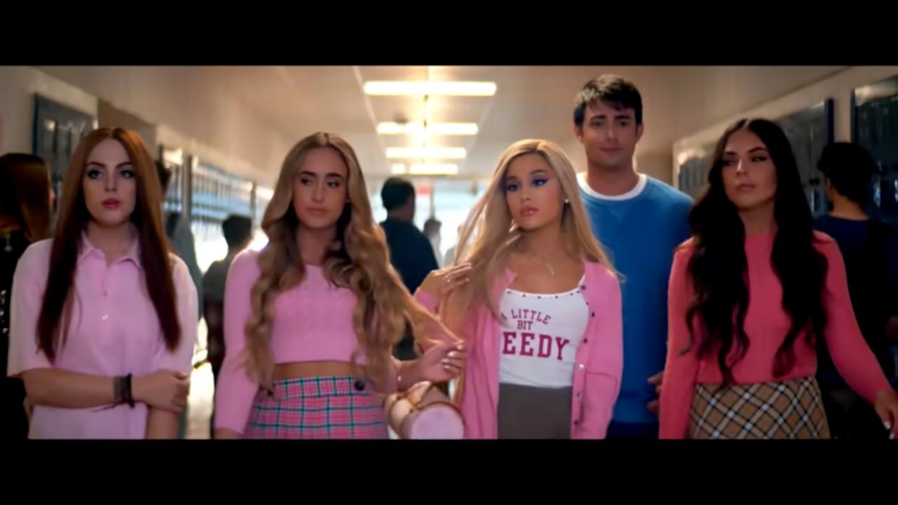 Still aus dem Musikvideo THANK U, NEXT von Ariana Grande: Vier junge Frauen und ein junger Mann, gekleidet im College-Stil, laufen in einem Schulgang nebeneinander auf die Kamera zu. Die Sängerin ist in der Mitte zu sehen.