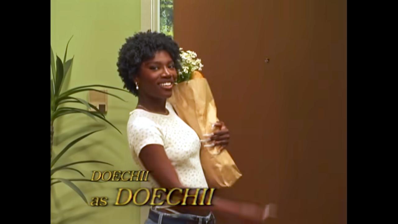 Still aus dem Musikvideo DENIAL IS A RIVER von Doechii: Die Sängerin Doechii betritt – wie in einer Sitcom – durch eine Tür einen Raum. Sie trägt eine Tüte mit Einkäufen im rm und lächelt direkt in die Kamera. Der Schriftzug "Doechii as Doechii" (Doechii als Doechii) ist eingeblendet.