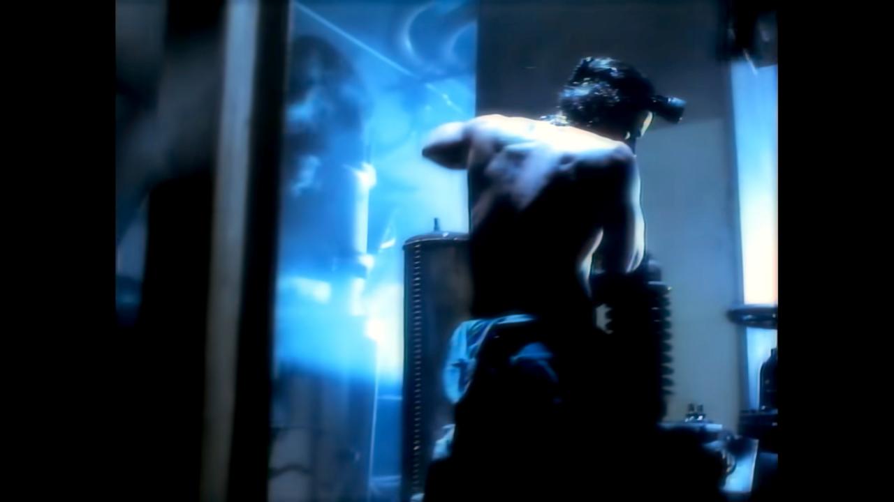 Still aus dem Musikvideo EXPRESS YOURSELF von Madonna: In einer industriellen, blau ausgeleuchteten Umgebung sieht man in der Rückenansicht einen muskulösen Arbeiter an einer Maschine.