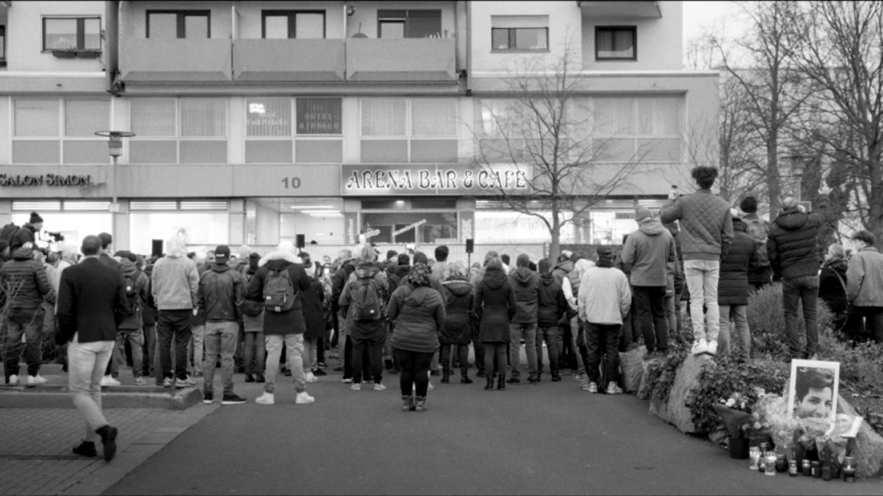 Still aus dem Dokumentarfilm DAS DEUTSCHE VOLK: Schwarz-Weiß-Aufnahme einer Gedenkfeier zum Anschlag von Hanau. Vor einem Tatort haben sich Menschen versammelt, die man in der Rückenansicht sieht.