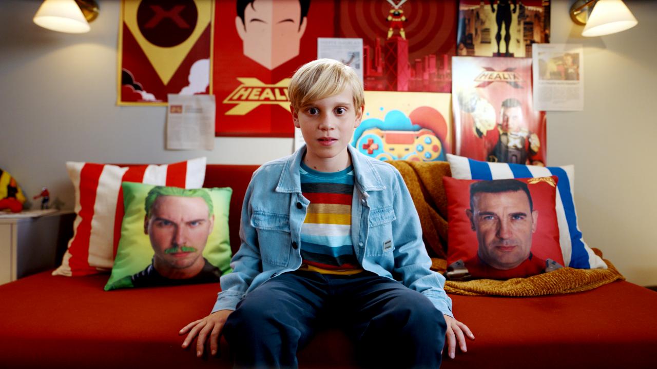 Still aus dem Kinderfilm SUPERKRÄFTE MIT KÖPFCHEN: Ein Junge sitzt auf einem Sofa und schaut direkt in die Kamera. Neben liegen zwei Kissen, auf denen die Bilder von zwei Männern abgedruckt sind.