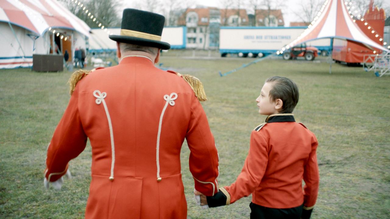 Still aus dem Dokumentarfilm ZIRKUSKIND: In der Rückenansicht: Ein Zirkusdirektor mit schwatzem Hut und roter Jacke und ein ebenfalls in rot gekleideter Junge laufen auf ein Zirkuszelt zu. Sie halten sich an den Händen fest. Der Junge schaut den älteren Herrn an.