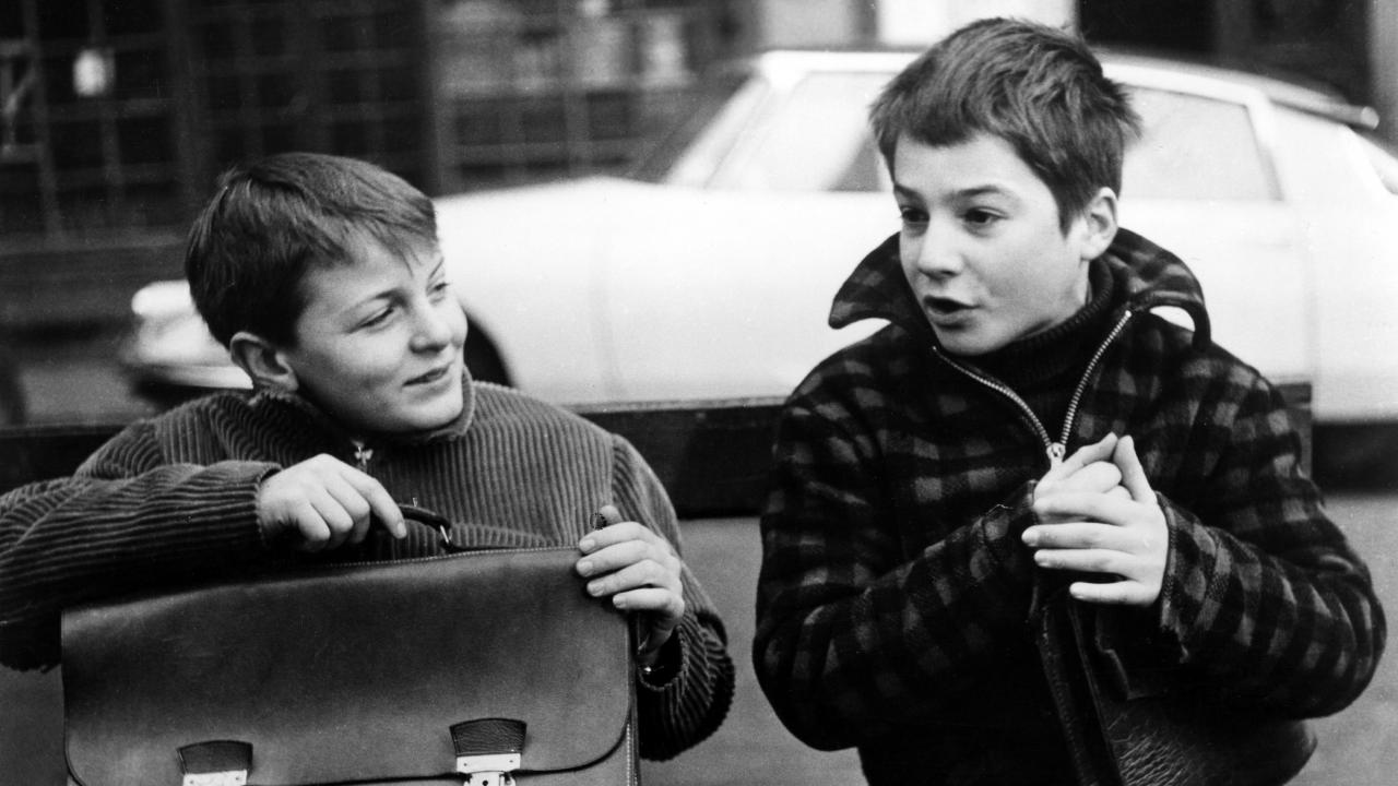 Still aus dem Spielfilm SIE KÜSSTEN UND SIE SCHLUGEN IHN: Zwei Jungen sitzen nebeneinander vor einer Straße. Der rechts Sitzende erzählt lehaft gestikulierend, der links von ihm hört sichtbar amüsiert zu.
