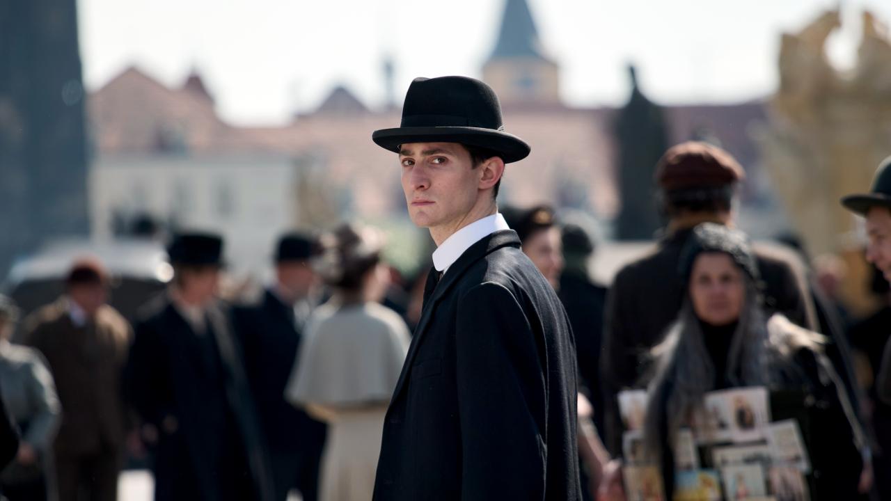 Filmstill aus dem Biopic FRANZ K.: Ein Mann, der dem Schriftsteller Franz Kafka ähnelt, dreht sich in einer Straßenszenerie um und schaut direkt in die Kamera. Um ihn herum sind Passanten zu sehen.