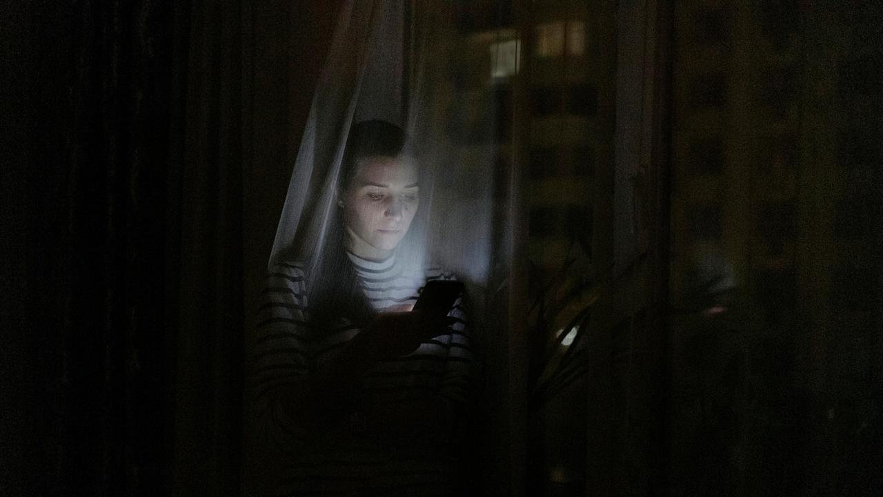 Still aus dem Dokumentarfilm SPLITTER AUS LICHT: Bild einer jungen Frau, die im Dunkeln hinter einem durchscheinenden Vorhang sitzt. Ihr Gesicht ist von einem Handy-Display erleuchtet.