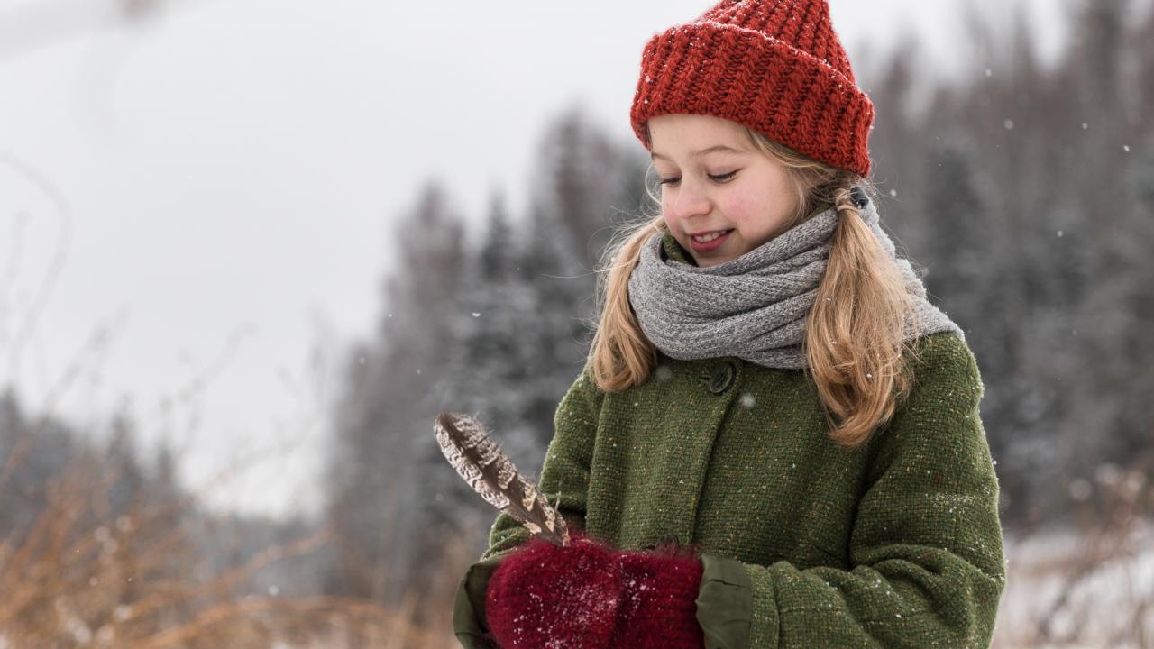 Still aus WEIHNACHTEN IM ZAUBEREULENWALD: Ein Mädchen steht in winterlicher Landschaft und hält die Feder eine Eule in der Hand, die sie lächelnd betrachtet.