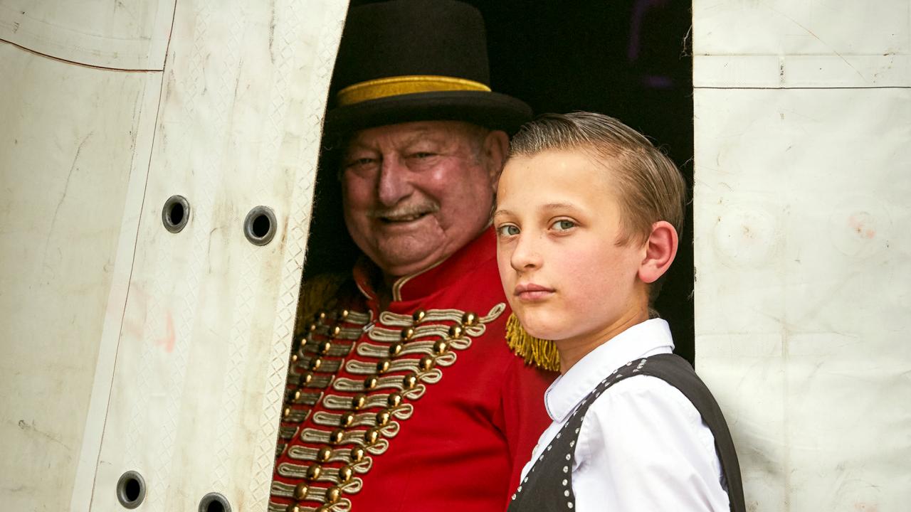 Still aus dem Dokumentarfilm ZIRKUSKIND: Eine Junge steht am Eingang eines Zirkuszelts und schaut direkt in die Kamera. Hinter ihm steht der Zirkusdirektor in roter Jacke.