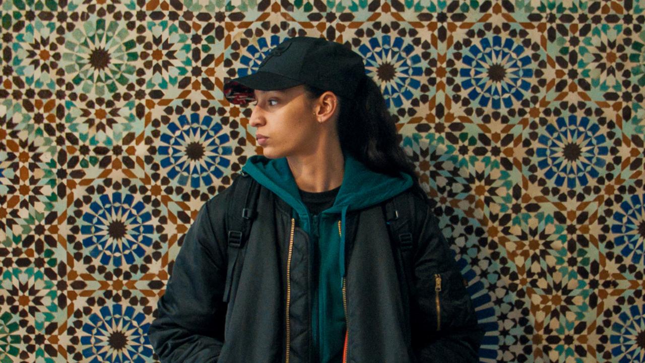 Filmstill aus dem Drama DIE JÜNGSTE TOCHTER: Eine junge, schwarz gekleidete Frau steht vor einer Wand mit bunt gemusterten Fliesen. Sie trägt eine Basecap und eine Bomberjacke und schaut ernst zur Seite.