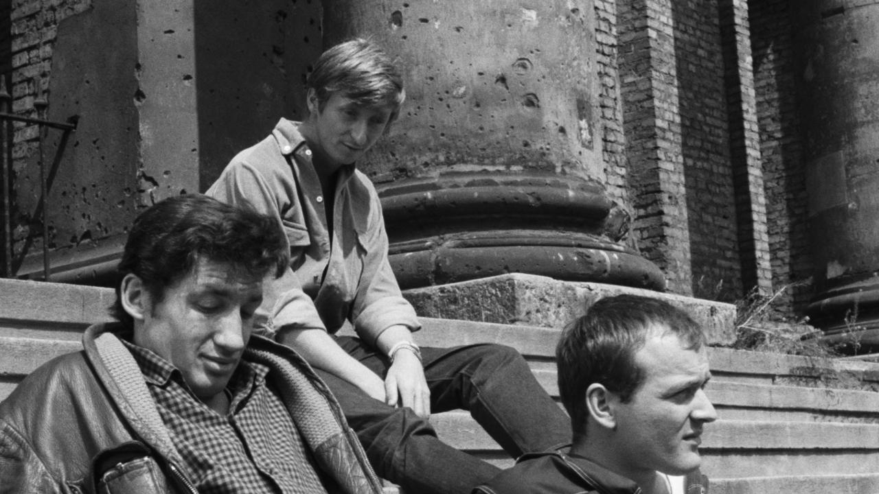 Filmstill aus dem DEFA-Film JAHRGANG 45: Drei junge Männer sitzen auf der Treppe eines Gebäudes, das Einschusslöcher hat.