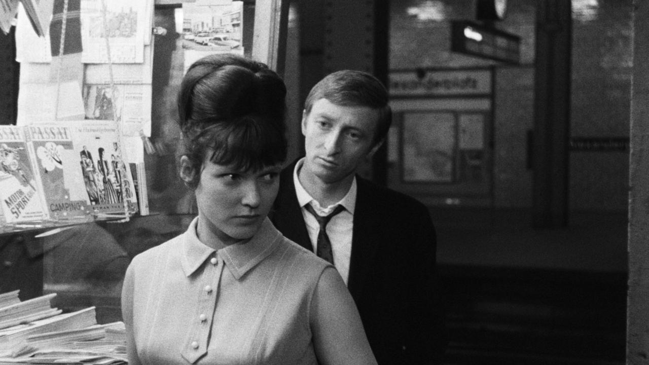 Filmstill aus dem DEFA-Film JAHRGANG 45: Eine junge Frau steht vor einem Geschäft und blickt leicht nach hinten. Hinter ihr steht ein junger Mann, der sie fragend anschaut.