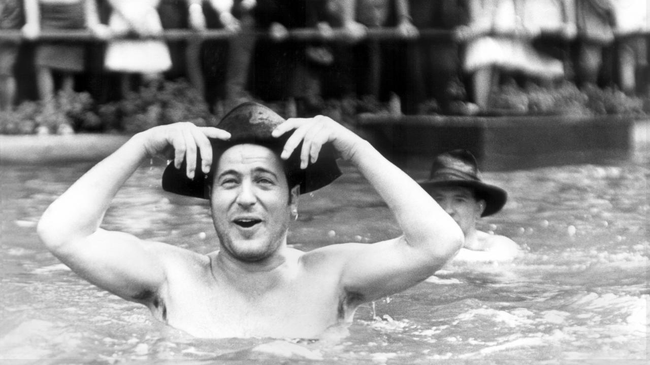 Filmstill aus SPUR DER STEINE: Ein Mann mit Zimmermannhut steht mit freiem Oberkörperin einem Brunnen, lacht und hält seinen Hut fest. Im Hintergrund sind ein weiterer Badender und Schaulustige zu sehen. 