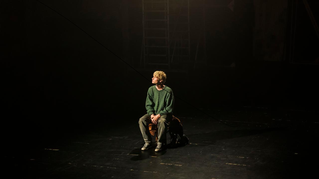 Filmstill aus ACH, DIESE LÜCKE, DIESE ENTSETZLICHE LÜCKE: Ein junger Mann sitzt allein auf einem Stuhl in einem leeren, dunklen Theatertaum.