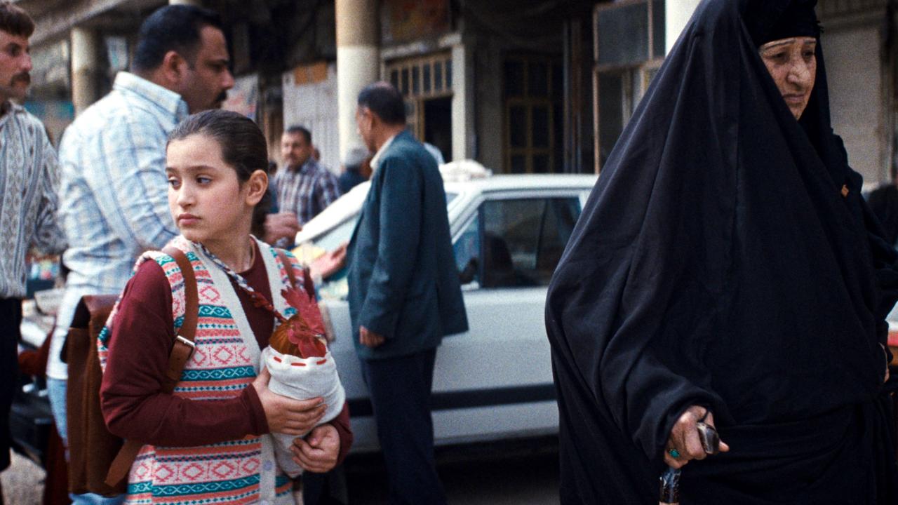 Filmstill aus EIN KUCHEN FÜR DEN PRÄSIDENTEN: Ein Schulmädchen, das eine Hahn in den Händen hält, läuft neben einer alten Frau, die in einen Tschador gekleidet ist, in einer Straße.