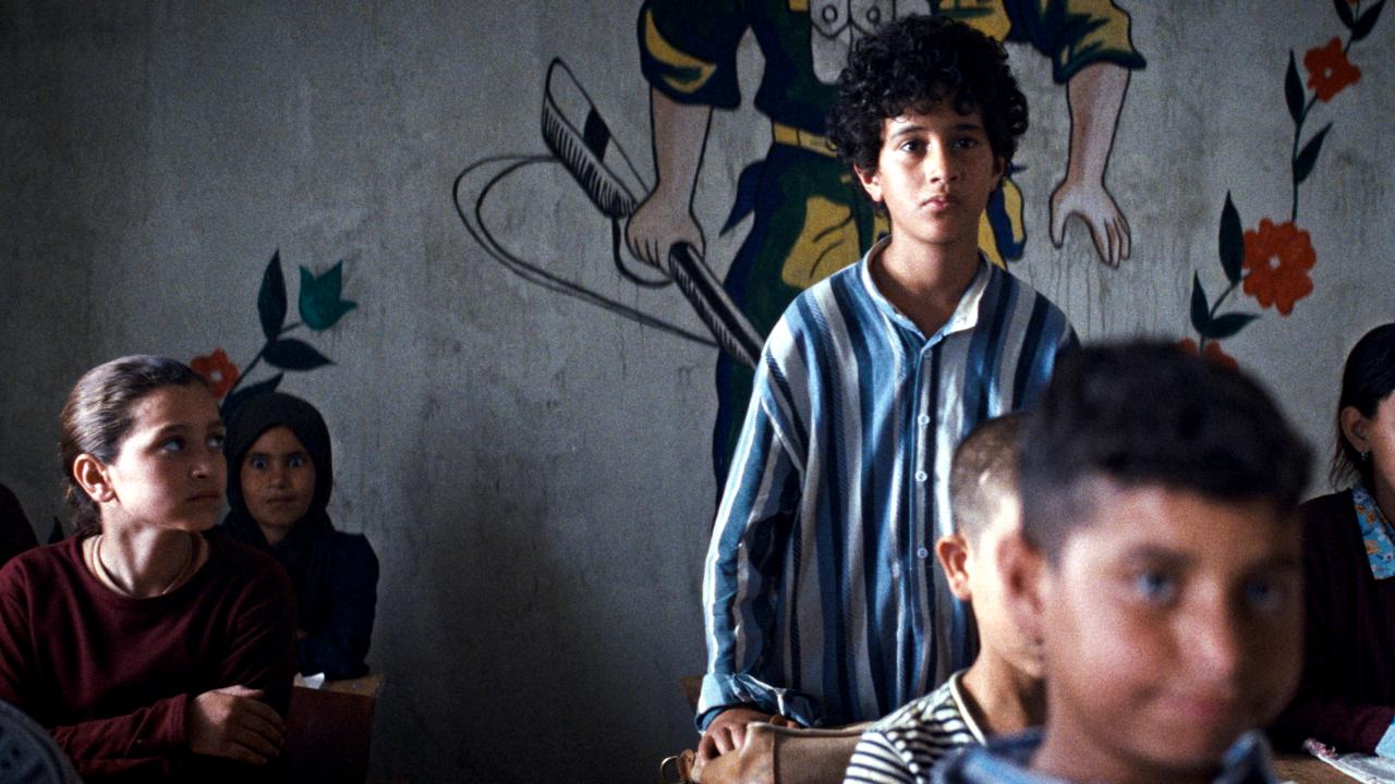 Filmstill aus EIN KUCHEN FÜR DEN PRÄSIDENTEN: In einer Schulklasse steht ein Junge, während alle anderen Kinder sitzen. Links neben sitzt ein Mädchen und schaut ihn an.